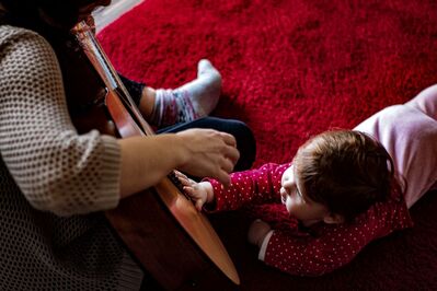 Baby lying on carpet patting guitar body as sitting adult plays the guitar.